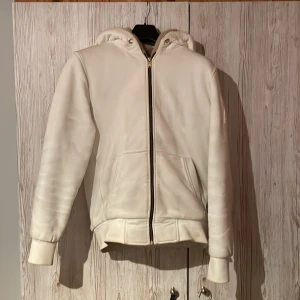 Vit hoodie från Moose Knuckles - Säljer en stilren vit hoodie från Moose Knuckles med guldfärgad logga på ärmen. Tröjan har huva, dragkedja framtill och två fickor. Insidan är svart med mönstrad lining. Perfekt för dig som gillar exklusiva streetwear-plagg. Saknar snören därav priset, använts en vinter går även att tvätta om man tar bort pälsen som går att ta bort