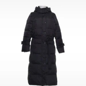 Svart lång pufferjacka med huva - Säljer en svart lång pufferjacka med huva och bälte i midjan. Jackan har knappar framtill, långa ärmar och quiltad design. Perfekt för kalla dagar när du vill hålla dig varm och samtidigt se cool ut.