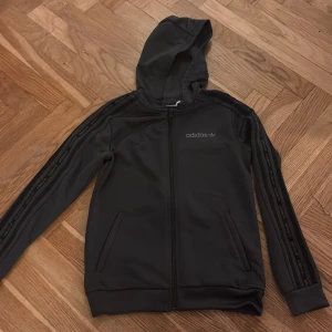 Svart hoodie från Adidas med huva - Svart hoodie från Adidas med klassiska ränder och logga längs ärmarna. Tröjan har huva, dragkedja framtill och två fickor. Materialet är mjukt och känns sportigt, perfekt för chill eller träning. Snygg och enkel design med diskreta detaljer.