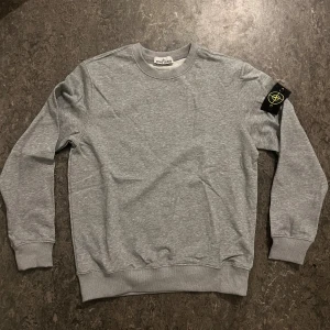 Grå sweatshirt från Stone Island - Snygg grå sweatshirt från Stone Island med klassisk rund hals och ribbade muddar. Tröjan har den ikoniska Stone Island-loggan på vänster ärm och är tillverkad i mjuk bomull. Perfekt för dig som gillar stilrena och bekväma plagg med streetkänsla.