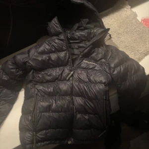 Svart Peak Performance dunjacka - Säljer en svart dunjacka från Peak Performance med huva och två dragkedjeförsedda fickor. Jackan är quiltad och har en snygg, puffig look som håller dig varm under kalla dagar. 