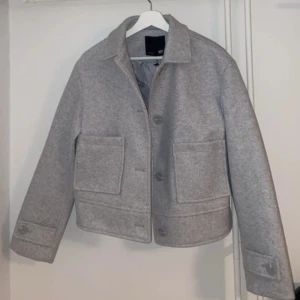 Grå ulljacka från 157 XS - Snygg grå jacka från 157 i storlek XS. Jackan har en rak, boxig passform med stora fickor framtill, klassisk krage och knäppning med knappar. Materialet är mjuk ullmix och insidan är fodrad för extra komfort. Perfekt för dig som gillar stilrena och enkla plagg.