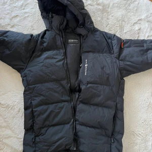 Svart dunjacka Sail Racing Antarctica - Svart dunjacka från Sail Racing Antarctica Expedition med huva och dragkedja. Jackan har logga på bröstet och ärmen, samt detaljerade broderier. Perfekt för kalla dagar med sin tjocka och isolerande design.