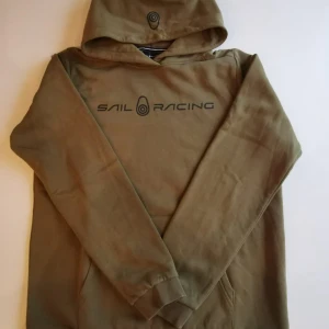 Olivgrön hoodie från Sail Racing - Olivgrön hoodie från Sail Racing med svart logga tryckt på bröstet och en mindre logga på huvan. Klassisk känguruficka framtill och ribbade muddar vid ärmslut och nederkant. Perfekt för en avslappnad och sportig stil.