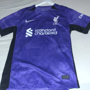 Lila Liverpool fotbollströja Nike S - Liverpool FC fotbollströja i lila med svarta detaljer, tillverkad i Dri-Fit material från Nike. Kortärmad med tryck från Standard Chartered och Expedia. Snyggt randigt mönster och klubbmärke på bröstet. Perfekt för dig som vill ha en unik supportertröja.