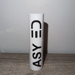 Vit ASYUYA Texture Powder - Säljer en vit ASYUYA Texture Powder i en cylinderformad plastflaska med svart text och en smiley på locket. Produkten har en praktisk pumpfunktion för enkel applicering. Perfekt för att ge håret extra volym och textur. Modern och stilren design.