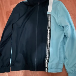 Lacoste blå och svart tracksuit - Säljer en snygg tracksuit från Lacoste i ljusblått och svart. Setet har dragkedja på jackan, elastiska muddar och coola Lacoste-loggor längs ena sidan. Materialet är lätt och passar perfekt för träning eller chill. Klassisk krokodil på byxan.