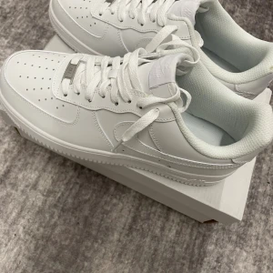 Nike Air Force 1 helvita sneakers - Fräscha Nike Air Force 1 sneakers i helvitt skinn med klassisk rund tå och platt sula. Ikonisk design med perforerade detaljer på tån, snörning och broderad Nike Air-logga på hälen. Perfekt för dig som gillar clean och stilren look.