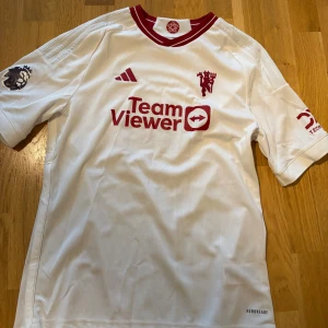 Manchester United vit fotbollströja - Säljer en vit Manchester United fotbollströja från Adidas med röda detaljer och logga. Tröjan har korta ärmar, rund hals med röd kant och Premier League-märke på ena ärmen. Materialet är lätt och andas, perfekt för match eller träning.