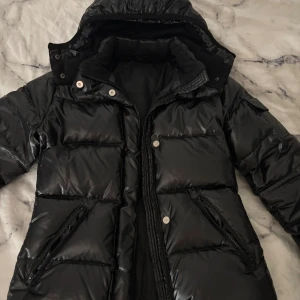 Svart pufferjacka med huva - Säljer en svart pufferjacka med glansig finish och huva. Jackan har dragkedja och knappar framtill, två fickor med dragkedja och en ficka med knapp på ärmen. Perfekt för kalla dagar och riktigt snygg till streetwear. Köptes för 2000kr.