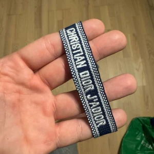 Blå Christian Dior J'Adior armband - Snyggt blått armband från Christian Dior med texten 'J'Adior' broderad i vitt. Armbandet har en bred, vävd design med vita detaljer längs kanterna och avslutas med flätade snören. Perfekt accessoar för att lyfta din outfit.