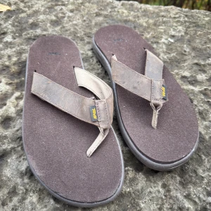 Bruna sandaler från Teva i skinn - Bruna flip-flop sandaler från Teva med breda remmar i mocka och mjuk fotbädd. Sulan är mönstrad för extra grepp och remmarna har en liten gul Teva-logga. Perfekta för varma dagar och enkel att ta på.