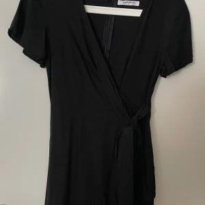 Svart omlottklänning Glamorous - Svart omlottklänning från Glamorous med korta ärmar och knyt i midjan. Klänningen har v-ringad hals och fint material.