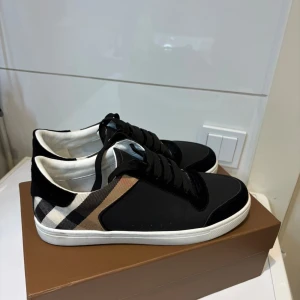 Burberry svarta sneakers med rutig detalj - Snygga svarta sneakers från Burberry med klassisk rutig panel i beige, svart och vit på sidorna. Skorna har rund tå, platt vit sula och svarta snören. Insidan är vit och materialet är en mix av skinn och textil. Perfekt för dig som gillar stilrena och lyxiga sneakers.