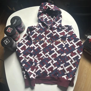 Mönstrad hoodie Tommy Hilfiger XS - Säljer en snygg hoodie från Tommy Hilfiger i storlek XS. Tröjan har ett stort mönster med vinröda, blå och vita färger samt logga broderad på bröstet. Klassisk huva med snörning och känguruficka framtill. Använd 5 gånger för 2 år sen och sedan dess bara legat i en garderob. Fet och unik för dig som vill sticka ut🤩                  Skick 9,5/10.      Pris kan diskuteras.