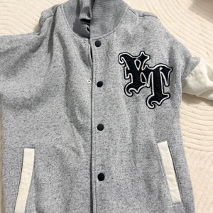 Grå bomberjacka med VT-tryck - Säljer en grå bomberjacka med vita detaljer och svart VT-tryck på bröstet. Jackan har ribbad krage, svarta knappar framtill och vita fickor. Perfekt för dig som gillar streetstyle och vill sticka ut med coola bokstäver.