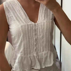 Vit blus med volang från La Cotière - Supersöt vit blus från La Cotière i storlek XS. Blusen har v-ringning, knappar framtill, spetsdetaljer och en volang längst ner. Ärmlös och tillverkad i mjukt bomullstyg, perfekt för varma dagar. Snygg att styla med jeans eller kjol!