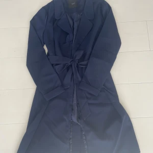 Mörkblå trenchcoat från Y.A.S - Snygg mörkblå trenchcoat från Y.A.S med klassisk krage och knytbälte i midjan. Jackan har lång ärm och är tillverkad i ett slätt, syntetiskt material med lite glans. Perfekt för dig som gillar stilrena och tidlösa plagg. Helt ny aldrig använd!
