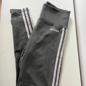 Grå Adidas leggings  - Snygga grå leggings från Adidas med klassiska vita ränder längs sidorna och logga framtill. Modellen är högmidjad och tillverkad i mjukt, stretchigt syntetmaterial med Aeroready-teknologi som andas. 