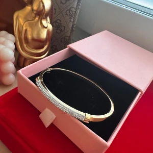 Guldfärgat armband med glittriga stenar - Snyggt guldfärgat armband med en rak, stilren form och en rad glittrande stenar framtill. Perfekt accessoar för att lyfta din outfit med lite extra bling. Kommer i en söt rosa ask.