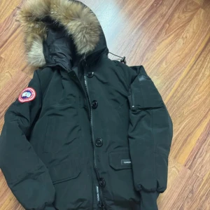 Svart dunjacka från Canada Goose - Säljer en svart dunjacka från Canada Goose med fet pälskant på huvan, stora fickor med lock och snygga detaljer som dragkedja och knappar framtill. Jackan har lång ärm och patch med logga på axeln. Perfekt för kalla vinterdagar och riktigt stilren.passar storlek S-M det är kort modell kolla bilder