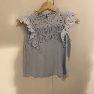 Ljusblå blus med spets från Holly & Whyte - Supersöt ljusblå blus från Holly & Whyte med volangdetaljer och broderad spets upptill. Blusen är ärmlös med högt halslinje och har fina hålmönster och rynkade partier framtill. Perfekt för dig som gillar romantisk och feminin stil.