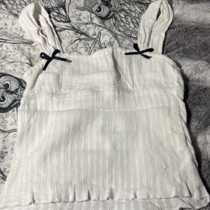 Vit linnetopp med rosetter från House - Supersöt vit linnetopp från House i storlek S. Toppen har breda axelband med små svarta rosetter framtill och är tillverkad i ett mjukt, strukturerat bomullstyg. Perfekt för varma dagar och har en enkel, stilren look med lätt rynkade detaljer.