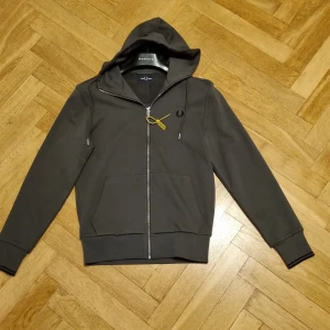 Mörkgrön hoodie från Fred Perry Ny, aldrig använd - Snygg mörkgrå hoodie från Fred Perry med dragkedja, huva och svarta detaljer. Tröjan har ribbade muddar med svarta ränder på ärmslutet och klassisk Fred Perry-logga broderad på bröstet. Perfekt för en chill och stilren look. Helt ny, aldrig använd då den tyvärr var för liten till sonen.