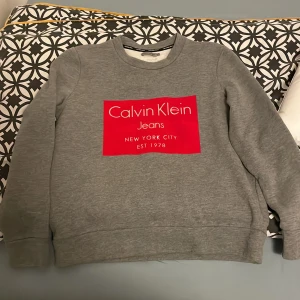 Grå sweatshirt från Calvin Klein Jeans - Säljer en grå sweatshirt från Calvin Klein Jeans med stor röd logga framtill. Tröjan har rund halsringning, långa ärmar och är tillverkad i mjuk bomullsblandning. Perfekt för en chill och stilren look. Skulle säga att den passar S för den är mindre i storleken.
