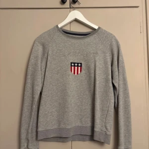 Grå sweatshirt från Gant med brodyr - Säljer en grå sweatshirt från Gant med broderad logga och sköld med stjärnor och ränder på bröstet. Tröjan har rund halsringning och långa ärmar, perfekt för en chill och sportig look.