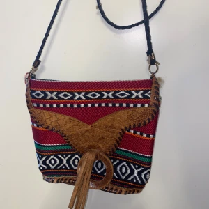 Färgglad axelväska med mönster och skinn - Unik axelväska med färgstarkt etnomönster i rött, svart, vitt, grönt och orange. Väskan har detaljer i brunt skinn, flätad axelrem och dekorativ frans framtill. Stängs med magnetknapp och har innerfack med dragkedja. Perfekt för dig som gillar boho-stil.
