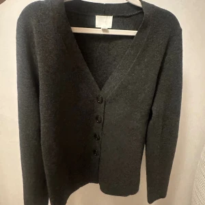 Mörkgrå kofta från H&M med knappar - Snygg mörkgrå kofta från H&M med v-ringad hals och fyra svarta knappar framtill. Koftan har långärmad design och är tillverkad i mjukt stickat material som känns skönt mot huden. Perfekt att slänga på sig när det är lite kyligt ute.