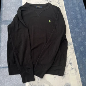 Svart sweatshirt från Polo Ralph Lauren - Svart långärmad sweatshirt från Polo Ralph Lauren med klassisk rund hals och grönt broderat logomärke på bröstet. Tröjan är i mjuk bomull och har ribbade muddar vid ärmslut och nederkant. Perfekt för chill dagar och enkel att matcha.
