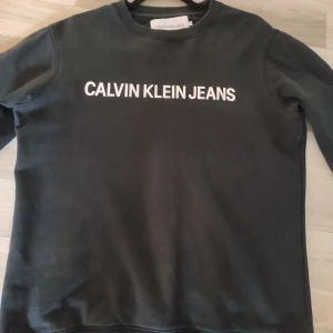 Svart sweatshirt från Calvin Klein Jeans - Svart sweatshirt från Calvin Klein Jeans med vit logotyp tryckt över bröstet. Tröjan har rund hals, långa ärmar och ribbade muddar vid ärmslut och nederkant. Perfekt för en clean och stilren look.