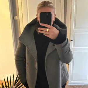Grå kappa - Snygg grå kappa med bred, uppvikt krage och asymmetrisk knäppning framtill. Jackan har en stilren look och är tillverkad i ett ulligt material som känns både mjukt och värmande. Perfekt för dig som gillar en klassisk men modern stil.