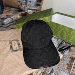 Svart Gucci keps med GG-mönster - Snygg svart keps från Gucci med klassiskt GG-monogram över hela kepsen. Justerbar rem bak i svart läder med gulddetalj. Kepsen har böjd skärm och är tillverkad i bomullsmaterial. Perfekt för dig som vill ha en lyxig och stilren accessoar.