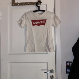 Vit Levi's t-shirt med röd logga - Vit t-shirt från Levi's med klassisk röd logga tryckt på bröstet. Rund hals och korta ärmar, tillverkad i mjuk bomull. Snygg och enkel design som funkar till jeans eller shorts. Perfekt för dig som gillar streetwear och ikoniska märken.