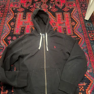 Svart hoodie från Polo Ralph Lauren - Svart hoodie från Polo Ralph Lauren med röd broderad logga på bröstet. Tröjan har vit snörning i huvan, dragkedja framtill och två stora fickor. Tillverkad i mjuk bomull, perfekt för chill dagar. Klassisk och stilren design som funkar året runt.           Skriv vid minsta funderingar😁l priset ej hugget i sten