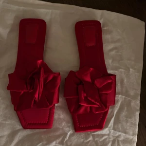 Röda sandaler med rosett från H&M - Snygga röda sandaler från H&M med fyrkantig tå och stora rosetter framtill. Skorna är tillverkade i satinliknande syntetmaterial och har platt sula. Perfekt för dig som vill sticka ut med färg och coola detaljer. Sandalerna är inte använda och är i nytt skick!