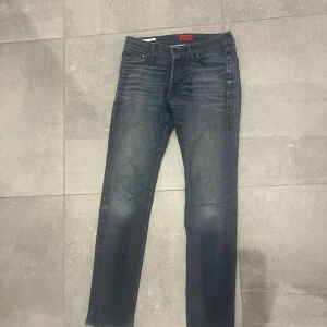 Slim fit jeans från Jack & Jones - Snygga slim fit jeans från Jack & Jones i mörkblå tvätt med coola slitningar och klassiska fem fickor. Jeansen har normal passform och raka ben, med detaljerade sömmar och knappstängning i midjan. Perfekt för dig som gillar en modern och stilren look. Storlek w30, l32