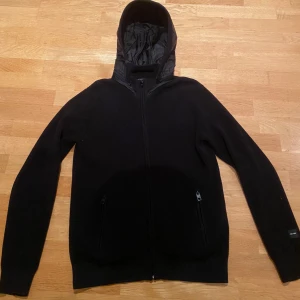 Svart stickad med dragkedja - Svart jacka med stickad kropp och quiltad huva. Jackan har hel dragkedja framtill och två fickor med dragkedja. Stilren design med diskret logga på ärmen, perfekt för dig som gillar enkel och clean look.