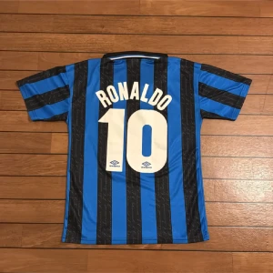 Inter Ronaldo #10 Umbro fotbollströja L - Retro Inter Milan fotbollströja från Umbro i storlek L, med Ronaldo och nummer 10 på ryggen. Tröjan har klassiska blå och svarta vertikala ränder, vit Pirelli-logga på bröstet och klubbmärke med stjärna. Tillverkad i lätt polyester med korta ärmar och krage.