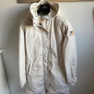 Beige parkajacka från Svea med huva - Parkajacka från Svea i beige med broderad amerikansk flagga på ärmen och huva med snörning. Jackan har knappar och dragkedja framtill, två fickor och broderad logga på bröstet. Perfekt för kalla dagar och har en clean, stilren look.