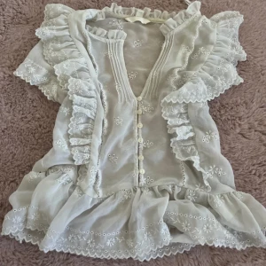 Vit blus med volanger och broderi - Supersöt vit blus med korta ärmar, volanger och broderade blommor. Blusen har knappar framtill, spetsdetaljer och v-ringad hals. Perfekt för dig som gillar romantisk och feminin stil. Materialet känns lätt och luftigt.