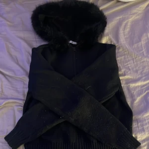 Svart pälsjacka med huva från Gina Tricot - Svart jacka från Gina Tricot med fluffig fuskpäls runt huvan och dragkedja framtill. Jackan har ribbade muddar och är långärmad, perfekt för kalla dagar. Stilen är både mysig och trendig, passar dig som gillar att sticka ut med detaljer.