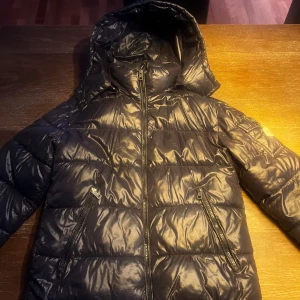 Svart Moncler pufferjacka med huva - Svart pufferjacka från Moncler med glansig finish, huva och dragkedja framtill. Jackan har två snedställda fickor med dragkedja och en ficka med Moncler-logga på ärmen. Pris kan diskuteras!