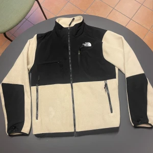  fleecejacka North Face - Snygg beige och svart fleecejacka från The North Face med hög krage och hel dragkedja. Jackan har tre fickor med dragkedja framtill och logga på bröstet och ryggen. Perfekt för dig som gillar stilren och funktionell design. Storlek M. Använd 3 gånger nästan helt ny. Nypris 2000