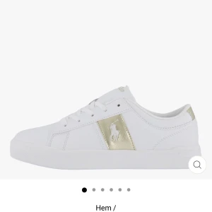 Vita sneakers från Polo Ralph Lauren - Fräscha vita sneakers från Polo Ralph Lauren med gulddetaljer och klassisk logga på sidan. Skorna har snörning, platt sula och är tillverkade i syntetmaterial. Perfekta för dig som gillar stilrena och sportiga vibes med en touch av lyx.