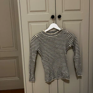 Randig långärmad tröja från Bellerose - Snygg långärmad tröja från Bellerose i beige och svart med klassiskt randigt mönster. Tröjan har en rund hals med lätt rynkad kant och är tillverkad i mjukt stickat material som sitter skönt mot kroppen. Perfekt för dig som gillar enkel och stilren design.