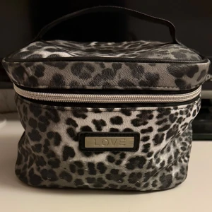 Leopardmönstrad handväska  - Snygg och praktisk handväska med leopardmönster i svart och grå toner. Väskan har en dragkedja runt locket och ett handtag upptill, perfekt för att bära med sig smink eller småprylar. Märke i metall framtill ger extra stilpoäng.
